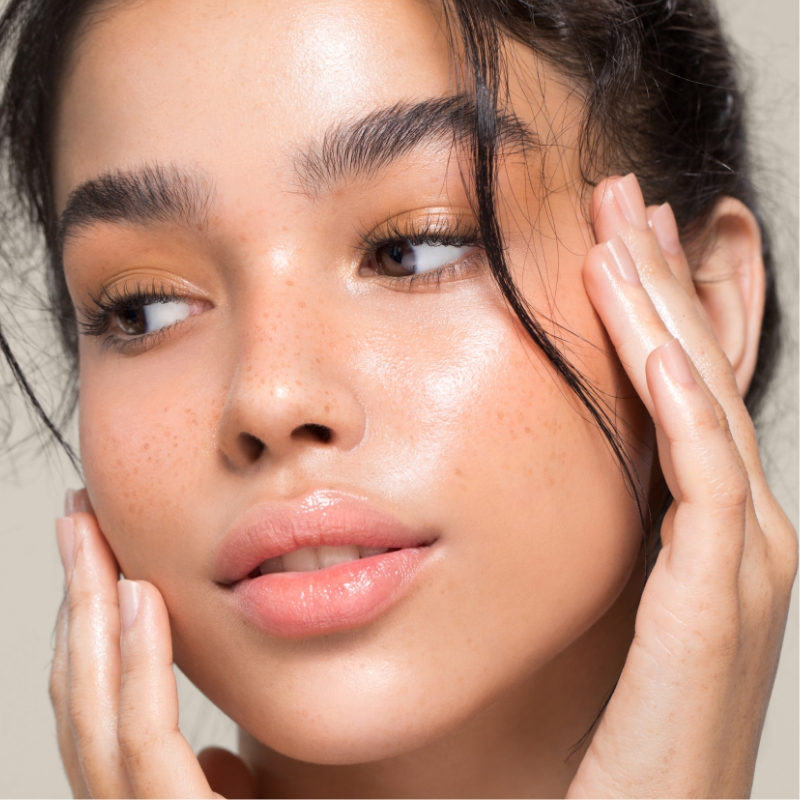 Woman with glowing skin and freckles gently touching her face.
